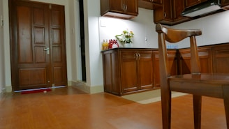 A warmly lit kitchen and entrance area featuring wooden cabinetry and flooring. A bouquet of flowers is placed on the kitchen counter next to several bottles. A wooden chair is in the foreground, and a closed door is visible on the left.