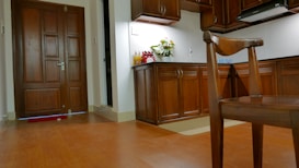 A warmly lit kitchen and entrance area featuring wooden cabinetry and flooring. A bouquet of flowers is placed on the kitchen counter next to several bottles. A wooden chair is in the foreground, and a closed door is visible on the left.