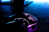 A sleek, modern gaming mouse with RGB lighting featuring blue and purple hues. The mouse is placed on a dark, textured surface, and has a unique, stylish design with a honeycomb pattern on the top.