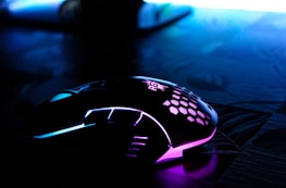 A sleek, modern gaming mouse with RGB lighting featuring blue and purple hues. The mouse is placed on a dark, textured surface, and has a unique, stylish design with a honeycomb pattern on the top.