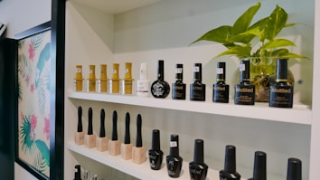 Shelves display an array of nail polish bottles arranged neatly. Various brands and shades are visible, including some with black, gold, and white labels. A small green houseplant adds a touch of nature to the setting, creating an inviting atmosphere. The background features a decorative floral pattern.