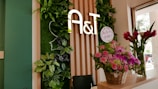 A welcoming interior space featuring a vertical garden with lush green leaves and a stylized outline of a face. The letters 'A&T' are prominently displayed on wooden paneling. A basket of pink and purple flowers is placed on a counter, alongside a tall vase with red flowers. Natural light streams in through a window, illuminating the scene.