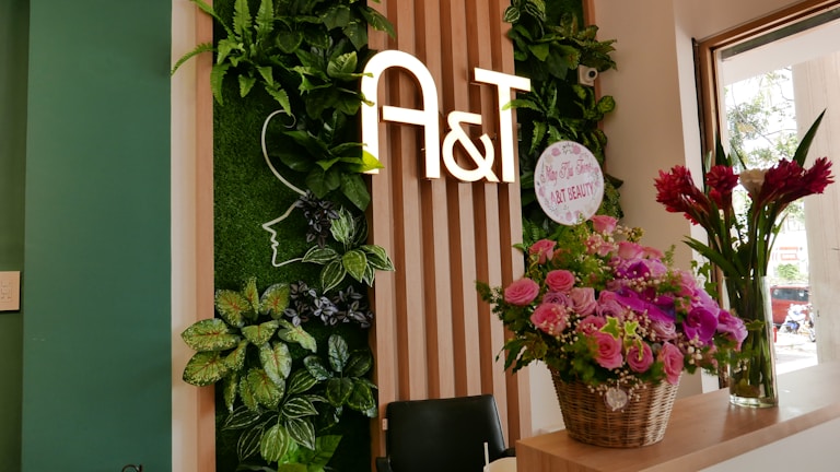 A welcoming interior space featuring a vertical garden with lush green leaves and a stylized outline of a face. The letters 'A&T' are prominently displayed on wooden paneling. A basket of pink and purple flowers is placed on a counter, alongside a tall vase with red flowers. Natural light streams in through a window, illuminating the scene.
