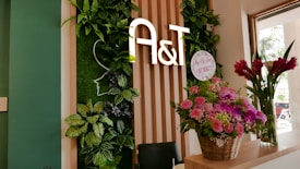 A welcoming interior space featuring a vertical garden with lush green leaves and a stylized outline of a face. The letters 'A&T' are prominently displayed on wooden paneling. A basket of pink and purple flowers is placed on a counter, alongside a tall vase with red flowers. Natural light streams in through a window, illuminating the scene.