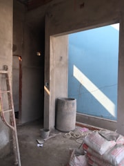A partially constructed room with unfinished cement walls, a metal ladder leaning against the left wall, and a large cylindrical container in the middle. Bags of construction material are stacked on the floor alongside loose ropes. Sunlight casts a diagonal shadow through an entrance, highlighting a blue-colored wall on the right.