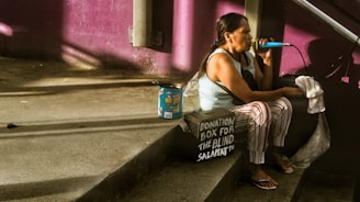 A woman sits on a concrete step, singing into a microphone. She has long dark hair tied back and is wearing a sleeveless top with striped pants. Next to her is a donation box labeled for the blind. The background features a purple wall, and sunlight casts shadows across the scene.