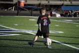 A young child on a football field is kicking a soccer ball. The child is wearing a black shirt with 'Lambert' and the number '2' in bright blue and red colors. The field markings are visible, and there's some seating in the background.
