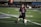 A young child on a football field is kicking a soccer ball. The child is wearing a black shirt with 'Lambert' and the number '2' in bright blue and red colors. The field markings are visible, and there's some seating in the background.