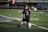 A young child on a football field is kicking a soccer ball. The child is wearing a black shirt with 'Lambert' and the number '2' in bright blue and red colors. The field markings are visible, and there's some seating in the background.