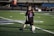 A young child on a football field is kicking a soccer ball. The child is wearing a black shirt with 'Lambert' and the number '2' in bright blue and red colors. The field markings are visible, and there's some seating in the background.
