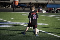 A young child on a football field is kicking a soccer ball. The child is wearing a black shirt with 'Lambert' and the number '2' in bright blue and red colors. The field markings are visible, and there's some seating in the background.