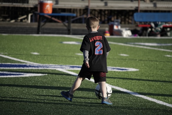 A young child on a football field is kicking a soccer ball. The child is wearing a black shirt with 'Lambert' and the number '2' in bright blue and red colors. The field markings are visible, and there's some seating in the background.