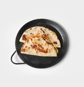 Two golden-brown quesadillas filled with melted cheese are placed on a round, black skillet. The quesadillas show slightly crispy edges and char marks, adding to their appetizing appearance. The background is a clean, white surface that contrasts with the dark skillet.