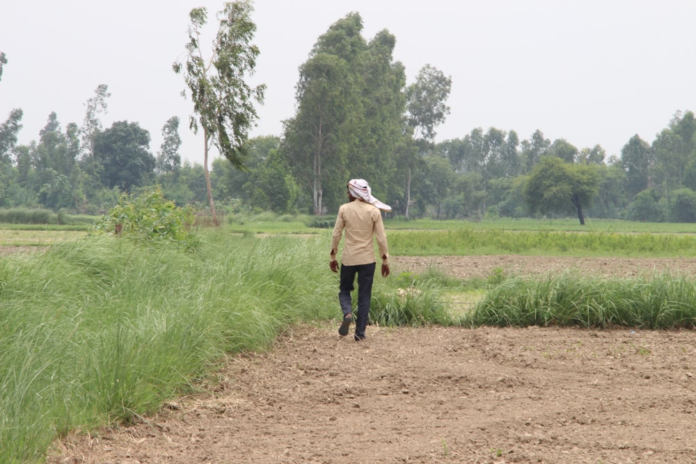 man in brown jacket walking on dirt road between green grass field during daytime