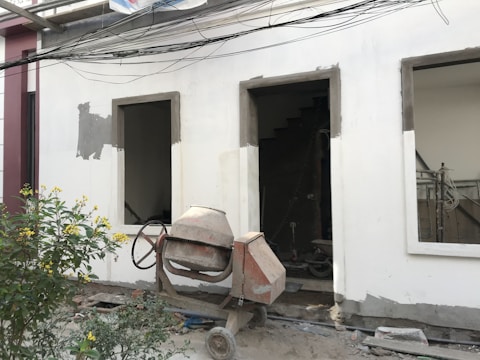 A construction site features an unfinished building facade with two rectangular open windows. In front of the building, a concrete mixer stands on the dusty ground, indicating ongoing construction work. A small bush with yellow flowers adds a touch of nature to the scene. Electrical wires run horizontally across the top portion of the image.
