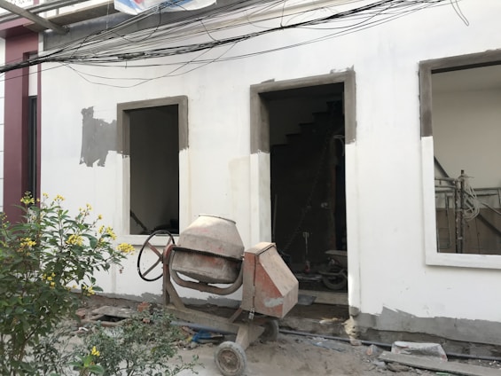 A construction site features an unfinished building facade with two rectangular open windows. In front of the building, a concrete mixer stands on the dusty ground, indicating ongoing construction work. A small bush with yellow flowers adds a touch of nature to the scene. Electrical wires run horizontally across the top portion of the image.