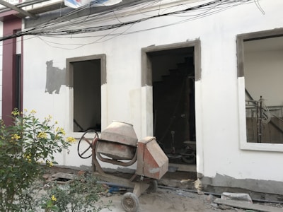 A construction site features an unfinished building facade with two rectangular open windows. In front of the building, a concrete mixer stands on the dusty ground, indicating ongoing construction work. A small bush with yellow flowers adds a touch of nature to the scene. Electrical wires run horizontally across the top portion of the image.