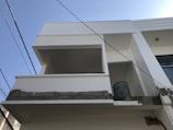 A modern white balcony under construction with visible electrical wires and a metal container. The building facade has a minimalist architectural design, with clean lines and geometric shapes.