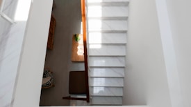 A top-down view of a modern interior space featuring a staircase on the right with white marble steps. To the left, there is a wooden table with a green plant in a pot and several glasses, next to a wicker chair with a patterned cushion. The space is well-lit with natural light.