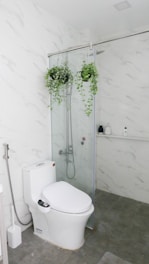 A modern bathroom with white marble-patterned walls and grey floor tiles. The space features a white toilet and a glass shower enclosure. Inside the shower are two hanging green plants, and various toiletries are visible on a small shelf.