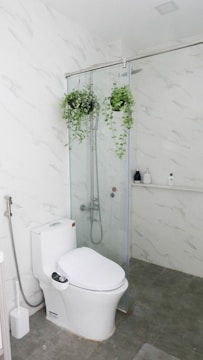 A modern bathroom with white marble-patterned walls and grey floor tiles. The space features a white toilet and a glass shower enclosure. Inside the shower are two hanging green plants, and various toiletries are visible on a small shelf.