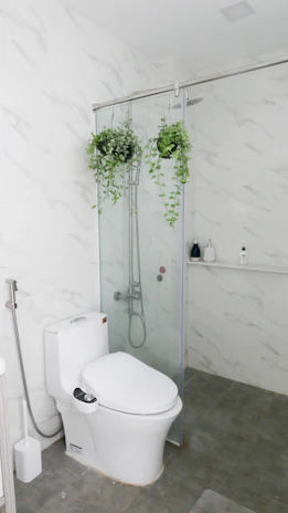 A modern bathroom with white marble-patterned walls and grey floor tiles. The space features a white toilet and a glass shower enclosure. Inside the shower are two hanging green plants, and various toiletries are visible on a small shelf.