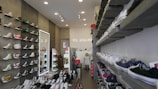 A well-organized shoe store with sneakers displayed on shelves along both walls. The left wall features high-top sneakers in various colors, while the right wall displays a variety of low-top sneakers. The store interior is well-lit with ceiling lights, and there are several rows of shoes placed on platforms in the center of the room. A mirror on the left wall reflects part of the store, and a sign with letters is visible in the background.