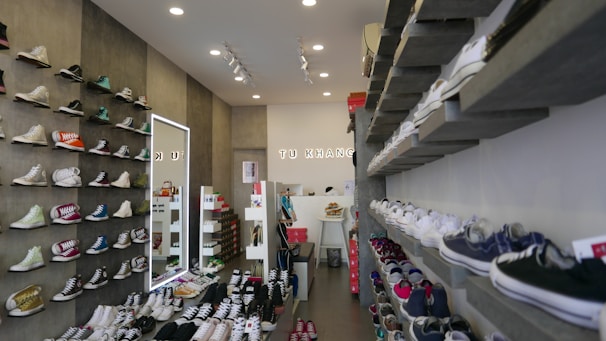 Brightly lit store interior showcasing rows of trendy men's sneakers and casual shoes neatly displayed on black and gold shelves.