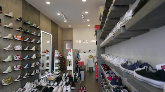 A well-organized shoe store with sneakers displayed on shelves along both walls. The left wall features high-top sneakers in various colors, while the right wall displays a variety of low-top sneakers. The store interior is well-lit with ceiling lights, and there are several rows of shoes placed on platforms in the center of the room. A mirror on the left wall reflects part of the store, and a sign with letters is visible in the background.