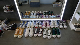 A variety of sneakers are neatly arranged in front of a large mirror on a store floor. The shoes are in several colors, including yellow, blue, white, pink, green, and grey. More shoes are reflected in the mirror, creating a symmetrical effect. The store has a clean and organized display, with shelves visible on the side.