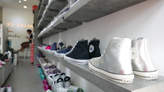 A shoe store with multiple shelves displaying a variety of sneakers. The shelves are arranged in a neat and organized manner with sneakers in different colors and styles. A person is seated at a counter in the background, possibly a store employee.