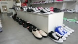 A shoe store display featuring a variety of sneakers arranged on a white platform and along the floor. The shoes include different styles and colors such as black, white, blue, and green. Shelves in the background hold additional shoes, and a sign indicates new arrivals.