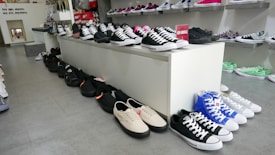 A shoe store display featuring a variety of sneakers arranged on a white platform and along the floor. The shoes include different styles and colors such as black, white, blue, and green. Shelves in the background hold additional shoes, and a sign indicates new arrivals.