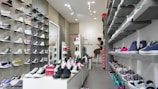 A shoe store interior with a variety of sneakers displayed on wall-mounted shelves on both sides. There are multiple colors and styles, including high-tops and low-tops. A counter at the back has a person seated, possibly assisting customers. A sign with 'NEW ARRIVALS' is visible on a central display table.