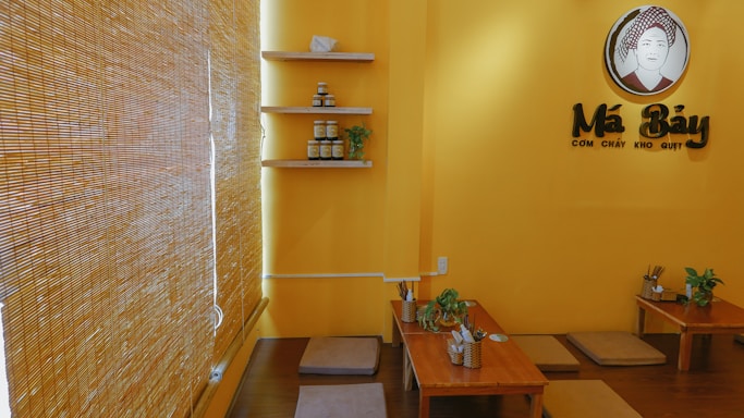 A cozy, warmly lit restaurant interior with yellow walls and bamboo blinds. There are low wooden tables with cushioned floor seating, decorated with small plants. Shelving on the wall holds jars and small decorative items. A sign featuring a stylized logo and text is prominently displayed.