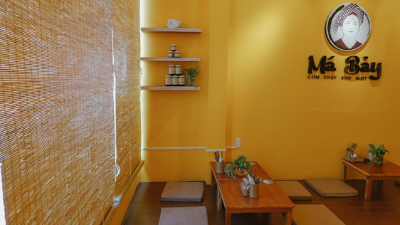 A cozy, warmly lit restaurant interior with yellow walls and bamboo blinds. There are low wooden tables with cushioned floor seating, decorated with small plants. Shelving on the wall holds jars and small decorative items. A sign featuring a stylized logo and text is prominently displayed.