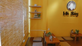 A cozy, warmly lit restaurant interior with yellow walls and bamboo blinds. There are low wooden tables with cushioned floor seating, decorated with small plants. Shelving on the wall holds jars and small decorative items. A sign featuring a stylized logo and text is prominently displayed.