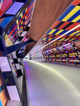 A modern pedestrian tunnel with a vibrant geometric pattern on the walls, featuring a combination of blue, red, yellow, orange, and black blocks. The ceiling has an LED strip running through the center, casting a white glow along the walkway, which has a metallic, textured surface.