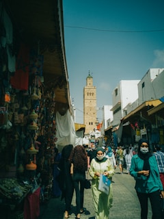 A bustling market scene with people walking along a narrow street. Stalls on either side are filled with colorful goods and items, while a tall, historical tower looms in the background against a clear blue sky. The atmosphere is vibrant, with a mix of shoppers and vendors.