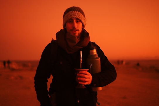 A person stands outdoors holding a cup with a metal straw, likely drinking mat&eacute;, and a thermos tucked under their arm. The person is dressed warmly in a beanie and scarf, and the sky has a vibrant, warm hue suggesting either sunrise or sunset.