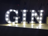 Large white letters spelling 'GIN' are illuminated with bright lights on a wooden surface against a dark background. The letters have an industrial or marquee-style design with individual lights embedded within each letter.