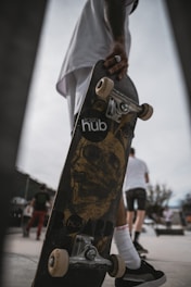 A skateboard is held by a person who is wearing dark sneakers and white socks. The skateboard features a graphic of a skull on the bottom, and the brand 'hub' is partially visible. In the background, several people are seen walking, and one appears to be wearing a white shirt and shorts.