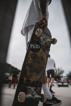 A skateboard is held by a person who is wearing dark sneakers and white socks. The skateboard features a graphic of a skull on the bottom, and the brand 'hub' is partially visible. In the background, several people are seen walking, and one appears to be wearing a white shirt and shorts.