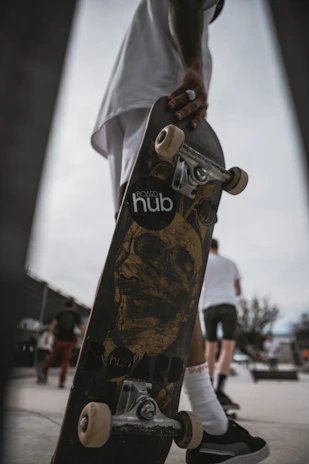 A skateboard is held by a person who is wearing dark sneakers and white socks. The skateboard features a graphic of a skull on the bottom, and the brand 'hub' is partially visible. In the background, several people are seen walking, and one appears to be wearing a white shirt and shorts.