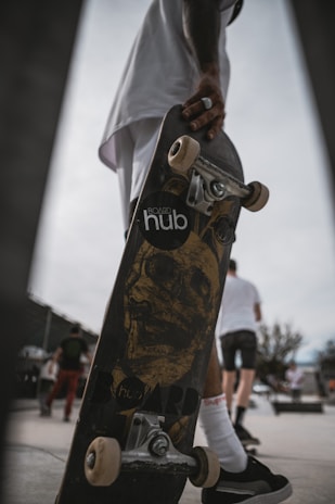 A skateboard is held by a person who is wearing dark sneakers and white socks. The skateboard features a graphic of a skull on the bottom, and the brand 'hub' is partially visible. In the background, several people are seen walking, and one appears to be wearing a white shirt and shorts.
