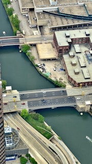 An aerial view of a city area featuring a river bordered by urban infrastructure. A multi-lane road bridge crosses the river, with visible cars traveling on it. Buildings with flat roofs and varied architectures surround the waterway. Green trees and patches of grass are scattered along the riverbanks, juxtaposed with concrete structures.