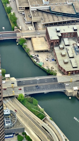 An aerial view of a city area featuring a river bordered by urban infrastructure. A multi-lane road bridge crosses the river, with visible cars traveling on it. Buildings with flat roofs and varied architectures surround the waterway. Green trees and patches of grass are scattered along the riverbanks, juxtaposed with concrete structures.