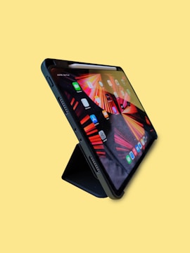 A tablet is positioned upright on a stand against a plain yellow background. The screen displays several colorful app icons and a vibrant abstract design with red and orange hues.