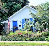 A charming white cottage with bright blue shutters and a matching door stands amidst a lush garden filled with colorful flowers and greenery. The front yard is adorned with a small black iron fence and an American flag is prominently displayed near the entrance. The vibrant garden includes daisies and other flowering plants.