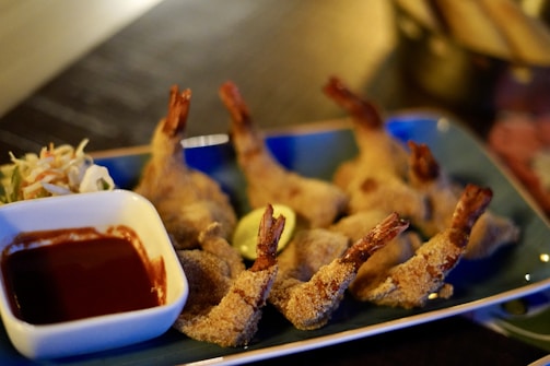 A plate of breaded shrimp is arranged in a row on a rectangular blue dish. Beside the shrimp is a small white bowl containing a dark red dipping sauce, possibly ketchup, and a serving of coleslaw garnished with shredded carrots. The presentation is neatly arranged with a warm, inviting aesthetic.